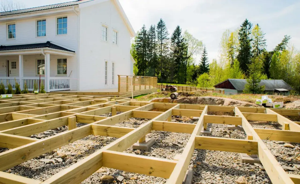 Grundl&auml;ggande stomme f&ouml;r altanen &auml;r p&aring; plats, redo att skruva trall. Ett vitt hus och gr&ouml;nskande tr&auml;d i bakgrunden.