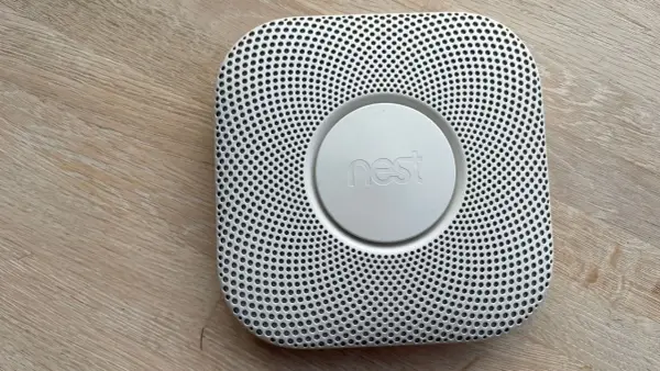 En vit Google Nest Protect-rökdetektor med ett runt "nest"-märke i mitten, placerad på ett trägolv.